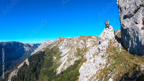 Couple with a hiking backpacks standing at the of a big boulder on the way to Hohe Weichsel in Austria, with a panoramic view on a vast valley. Narrow pathway. They are enjoying the view, having fun