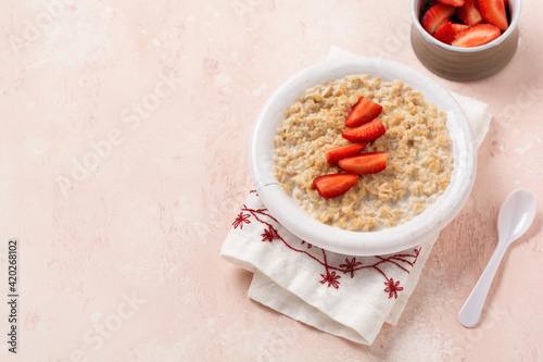 Simple oatmeal porridge with strawberries in a white plate on a linen napkin on pink background. Breakfast health food concept. Top view
