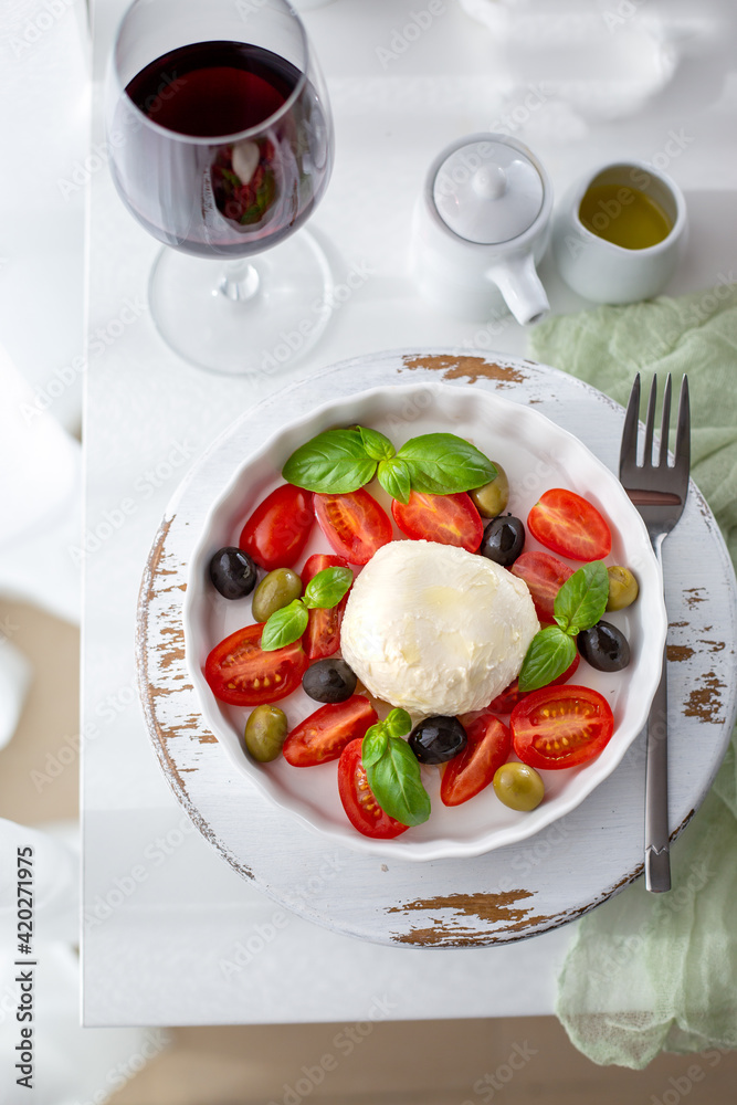 Mozzarella cheese salad plate with fresh tomatoes, black and green olives, basil. A glass of red wine, olive oil dressing.