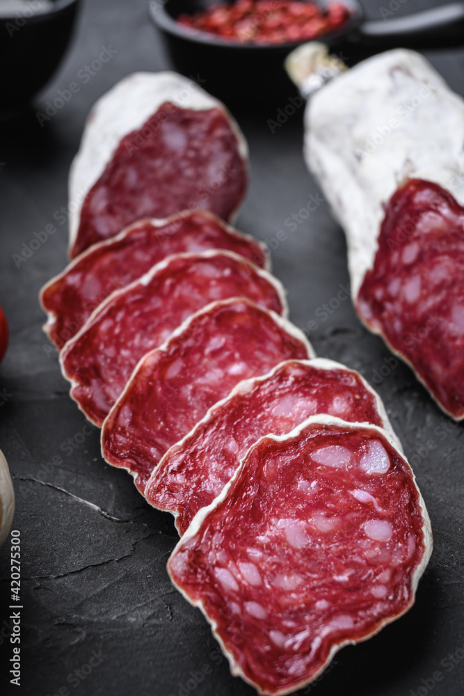 Dry cured salchichon sausage slices with herbs on black textured background