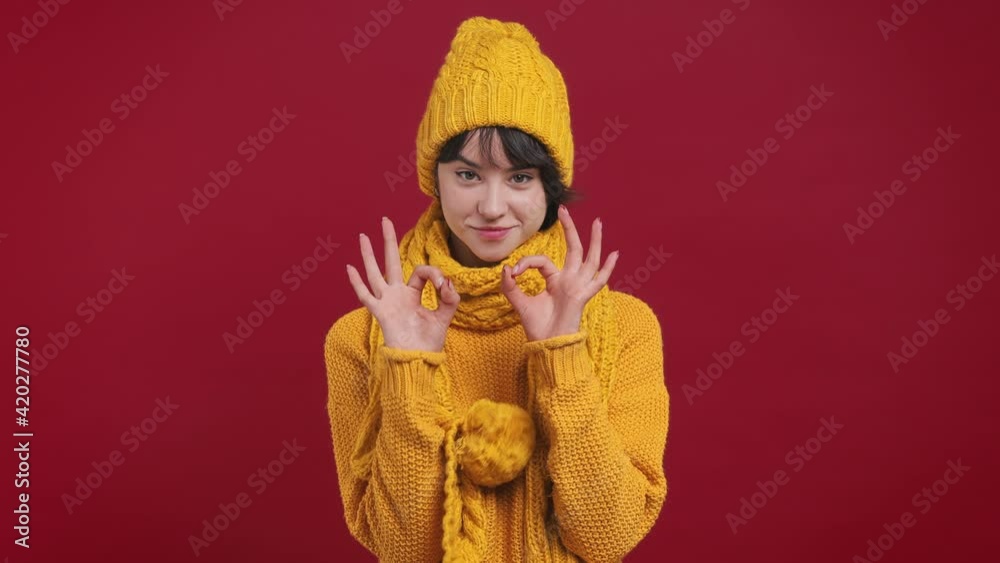 Smiling charming fun cheerful short hairdo young woman 20s years old in yellow knitted sweater hat scarf look camera showing okay ok zero fingers gesture isolated on bright vivid red background studio Smiling charming fun cheerful short hairdo young woman 20s years old in yellow knitted sweater hat scarf look camera showing okay ok zero fingers gesture isolated on bright vivid red background studio