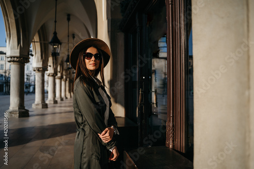 Outdoor fashion portrait of elegant, luxury woman wearing beige hat, sunglasses, trendy white shirt, in a green trench coat, walking in street. Copy, empty space for text