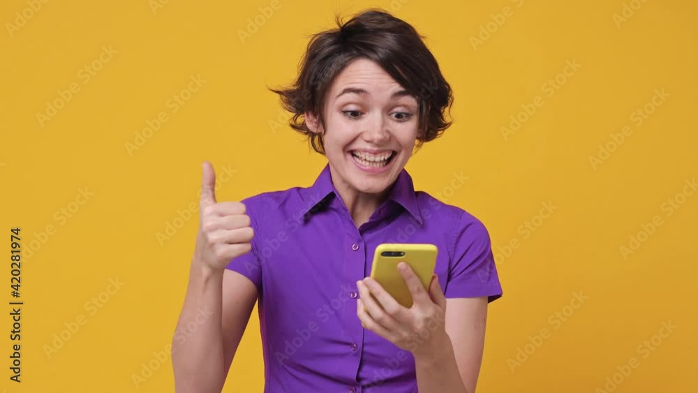 Happy excited surprised young woman in violet shirt hold using mobile cell phone typing browsing say wow yes just found out great big win news pointing on gadget isolated on yellow background studio Happy excited surprised young woman in violet shirt hold using mobile cell phone typing browsing say wow yes just found out great big win news pointing on gadget isolated on yellow background studio