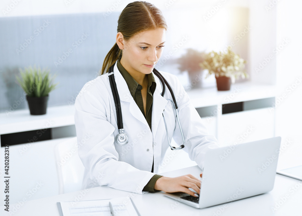 Woman-doctor typing on laptop computer while sitting in sunny hospital office. Data in medicine and healthcare Woman-doctor typing on laptop computer while sitting in sunny hospital office. Data in medicine and healthcare