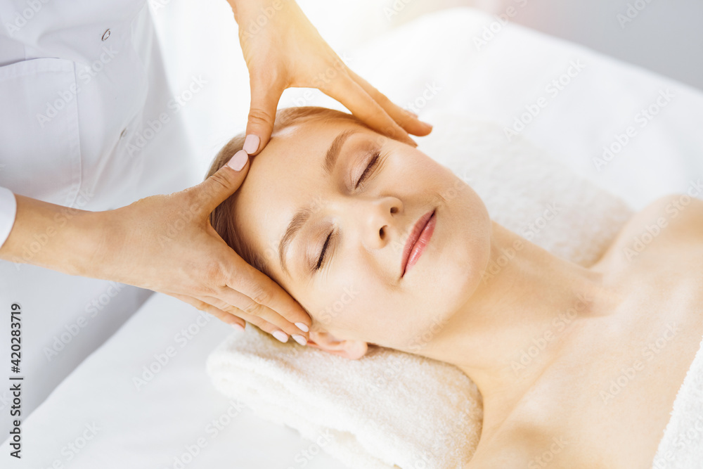 Beautiful caucasian woman enjoying facial massage with closed eyes in sunny spa salon. Relaxing treatment in medicine Beautiful caucasian woman enjoying facial massage with closed eyes in sunny spa salon. Relaxing treatment in medicine