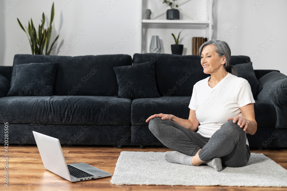 Smiling and active senior woman sits on the floor in lotus pose, looks online yoga tutorial on the laptop, elderly female practicing yoga at home with video classes, online training, healthy lifestyle