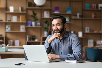 Smiling bearded indian businessman working on laptop in modern office lobby space. Young indian student using computer remote studying, watching online webinar, virtual training on video call meeting.