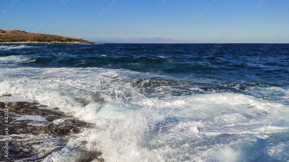 Sunny clear stormy blue sea hitting rocky shore. Summer sea with white waves on Aegean sea in Athens, Greece