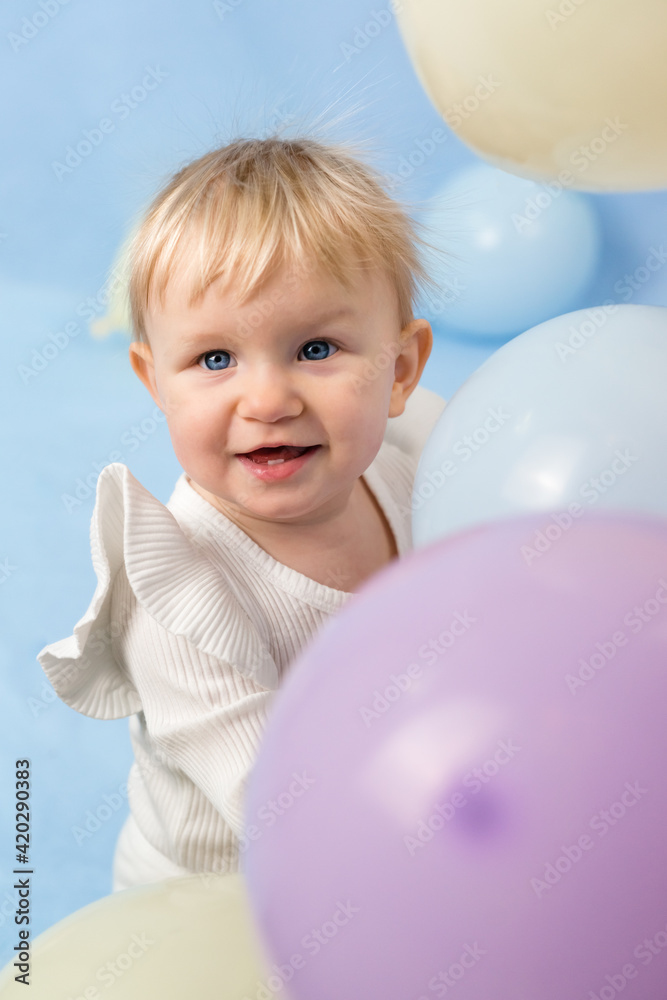 Beautiful cheerful little girl 1 year old with blue eyes in white turtleneck among balloons and looks at the camera Beautiful cheerful little girl 1 year old with blue eyes in white turtleneck among balloons and looks at the camera