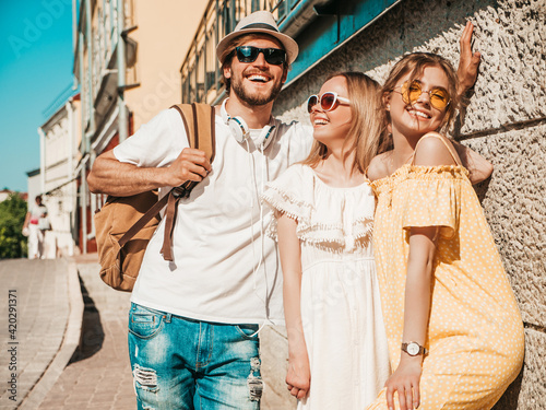 Group of young three stylish friends posing in the street. Fashion man and two cute female dressed in casual summer clothes. Smiling models having fun in sunglasses.Cheerful women and guy outdoors