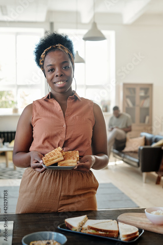 Young happy African female with homemade waffles looking at you while standing by table