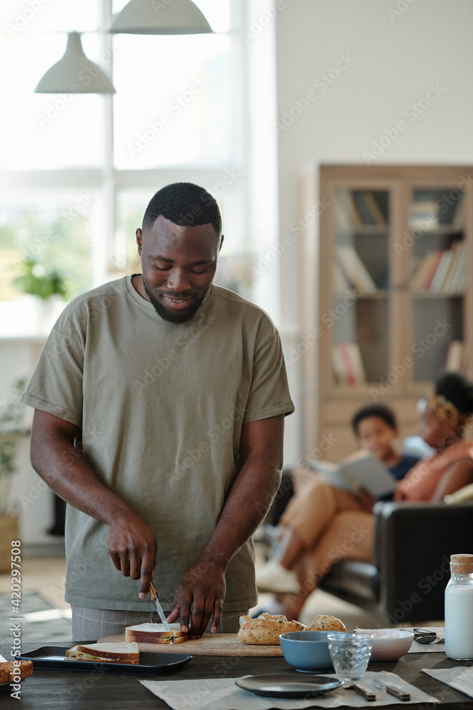 Young African man in casualwear preparing sandwiches for breakfast by table in the kitchen