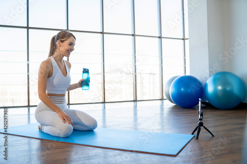 Cheerful sports girl sits in front of phone camera and records online workout. Slender woman in white sportswear sits on yoga mat and conducts an internet stream for subscribers