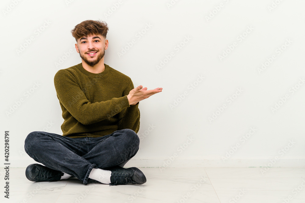 Young Moroccan man sitting on the floor isolated on white background holding a copy space on a palm.