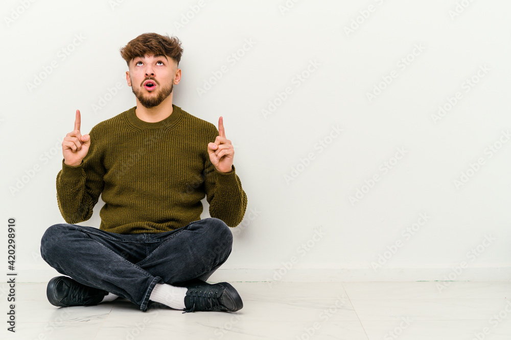 Young Moroccan man sitting on the floor isolated on white background pointing upside with opened mouth.