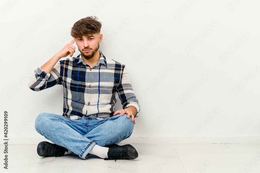 Young Moroccan man sitting on the floor isolated on white background pointing temple with finger, thinking, focused on a task. Young Moroccan man sitting on the floor isolated on white background pointing temple with finger, thinking, focused on a task.