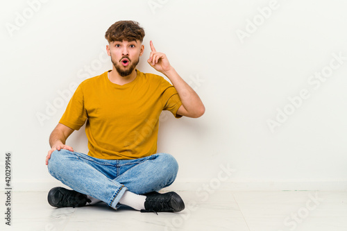 Young Moroccan man sitting on the floor isolated on white background pointing upside with opened mouth. © Asier