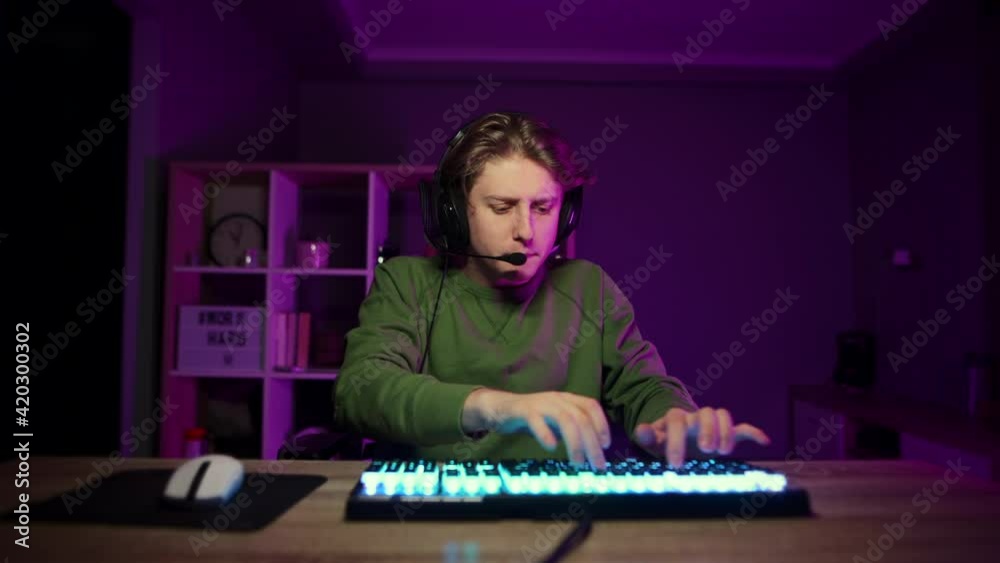Emotional guy in a headset plays online games and streaming. Focused gamer with a tense face plays video games at home on a computer in a room with purple lights and angrily looks at the camera. Emotional guy in a headset plays online games and streaming. Focused gamer with a tense face plays video games at home on a computer in a room with purple lights and angrily looks at the camera.