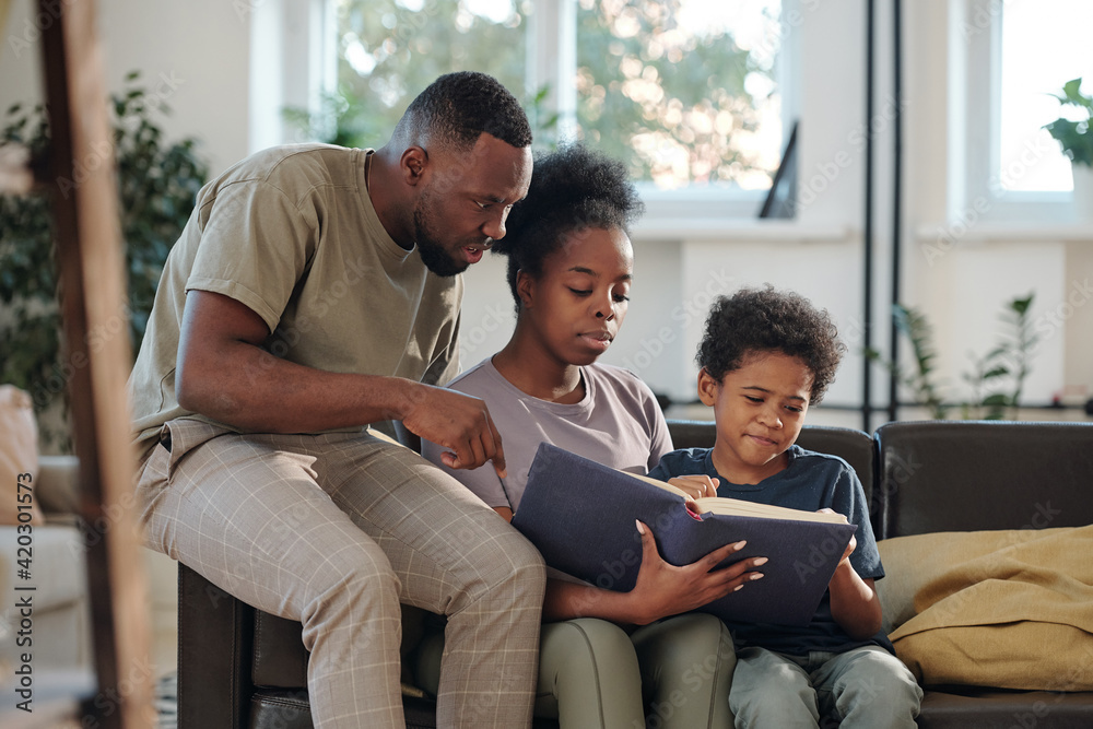 Young African family of parents and cute little son reading large book of tales or encyclopaedia Young African family of parents and cute little son reading large book of tales or encyclopaedia