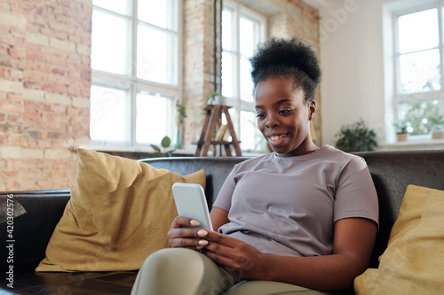 Restful young African woman with smartphone sitting on black leather couch and watching funny video