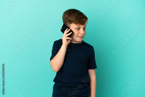 Little redhead boy isolated on blue background keeping a conversation with the mobile phone