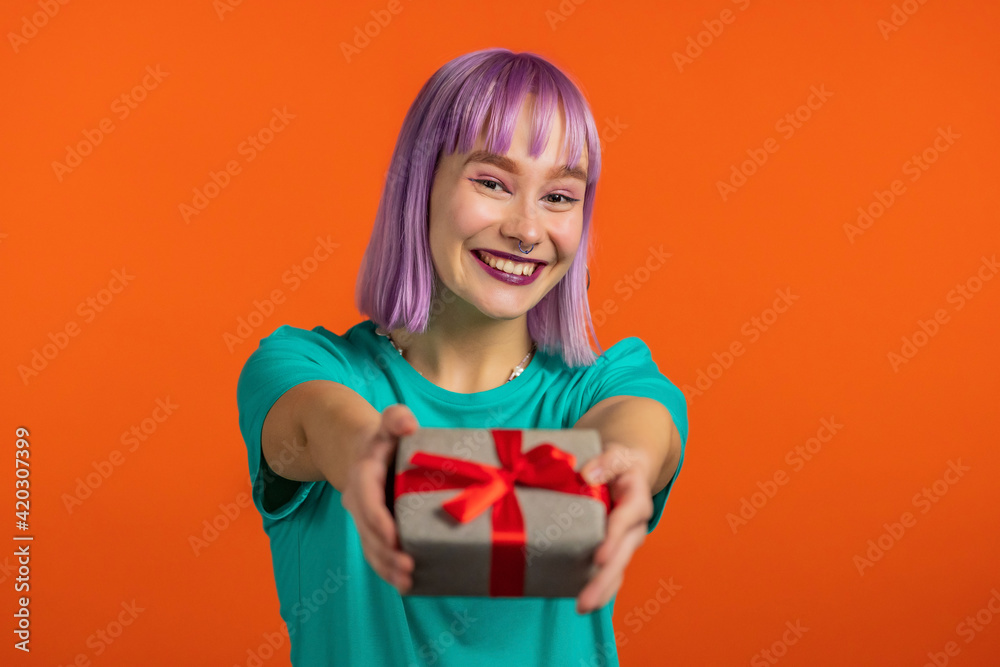 Excited woman with violet hair holding gift box and gives it by hands to camera on orange wall background. Girl smiling, she is happy with present. Studio portrait Excited woman with violet hair holding gift box and gives it by hands to camera on orange wall background. Girl smiling, she is happy with present. Studio portrait