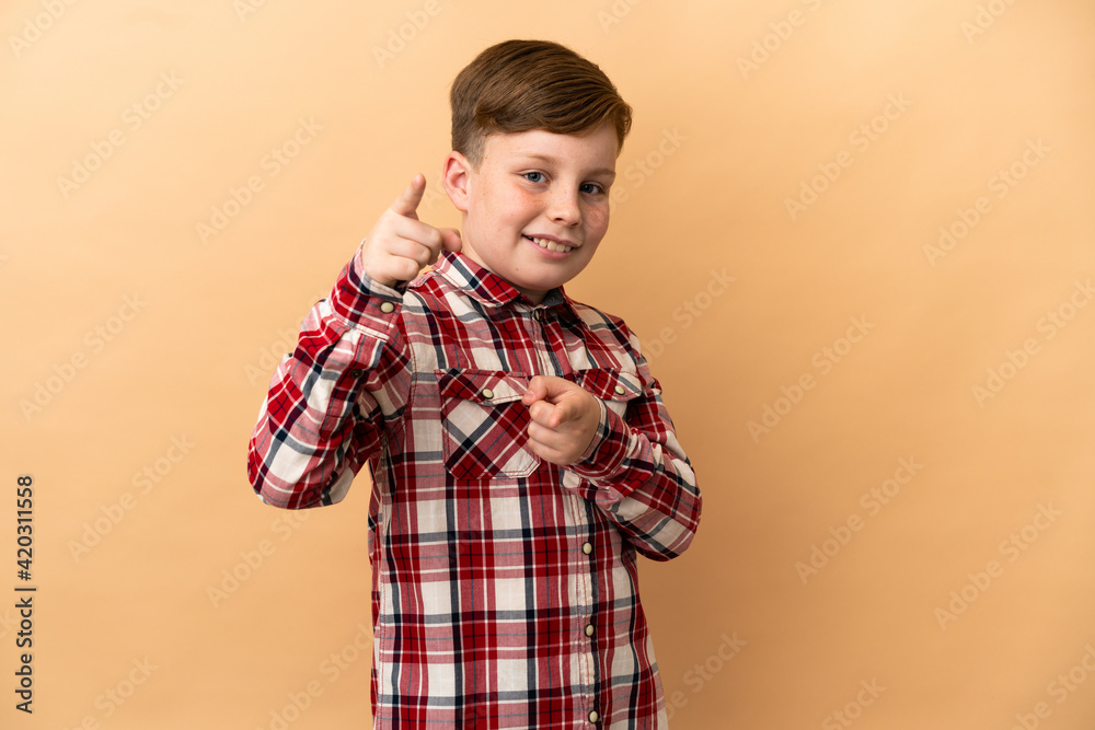 Little redhead boy isolated on beige background points finger at you with a confident expression Little redhead boy isolated on beige background points finger at you with a confident expression