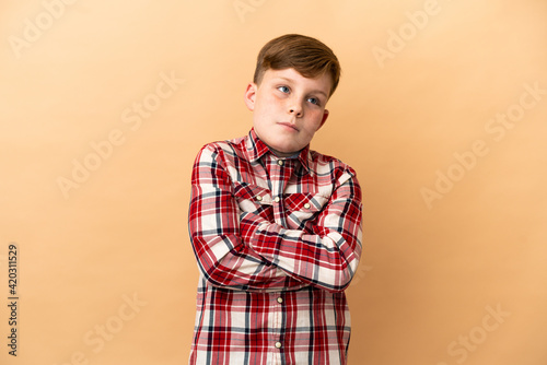 Little redhead boy isolated on beige background making doubts gesture while lifting the shoulders
