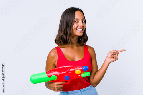 Young mixed race woman holding a water gun isolated smiling cheerfully pointing with forefinger away.