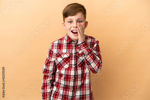 Little redhead boy isolated on beige background shouting with mouth wide open