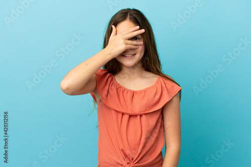 Little caucasian girl isolated on blue background covering eyes by hands and smiling