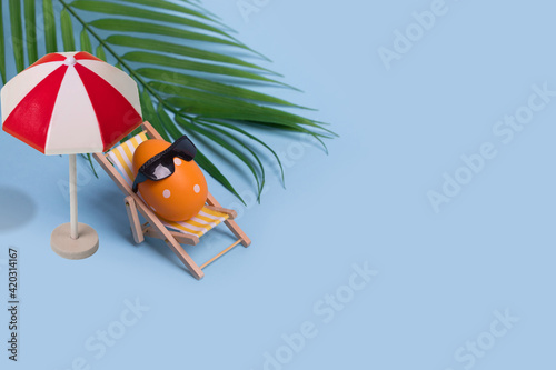 Fotografia Creative funny composition with Easter egg with sunglasses sitting on deck chair and sun umbrella on bright blue background