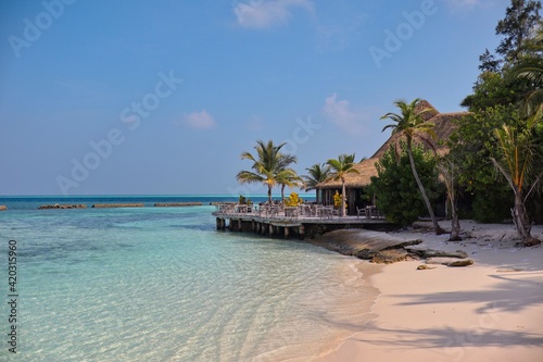Fototapeta Naklejka Na Ścianę i Meble -  Maldives Resort with Sandy Beach, Turquoise Sea and Thatched Restaurant. Sunny Day in Paradise. Maldivian Komandoo Island Resort.