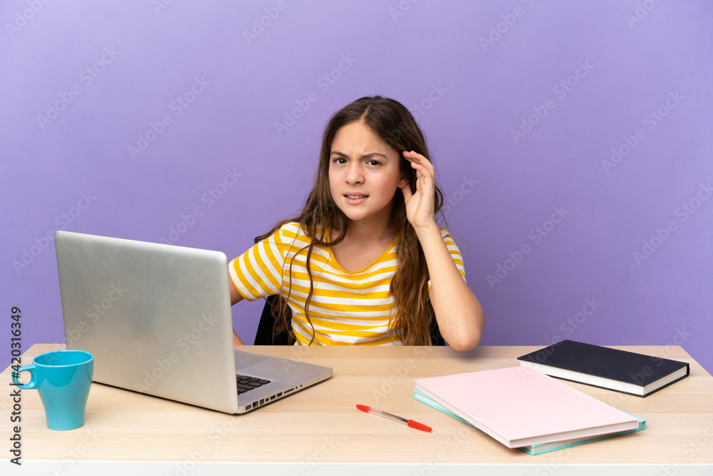 Little student girl in a workplace with a laptop isolated on purple background listening to something by putting hand on the ear Little student girl in a workplace with a laptop isolated on purple background listening to something by putting hand on the ear