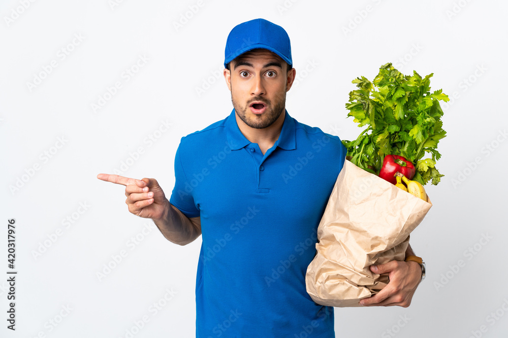 Delivery man with vegetables isolated on white background surprised and pointing side Delivery man with vegetables isolated on white background surprised and pointing side