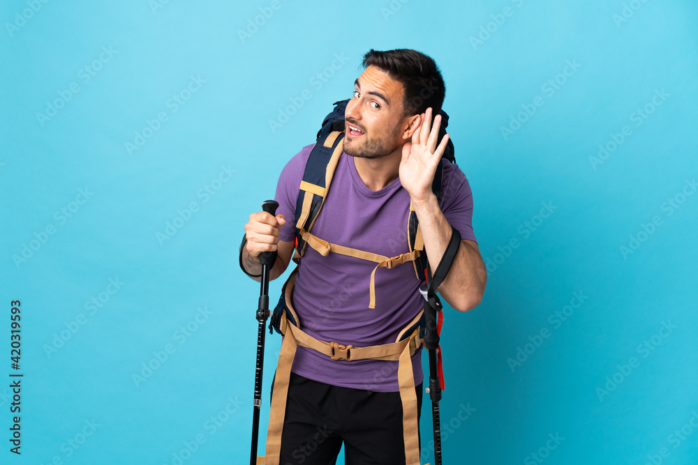Young caucasian man with backpack and trekking poles isolated on blue background listening to something by putting hand on the ear