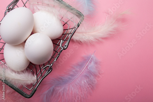 White Easter eggs in a grocery basket, shopping cart, multicolored delicate bird feathers on a pink background