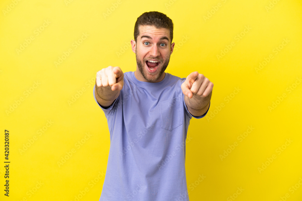 Handsome blonde man over isolated yellow background surprised and pointing front Handsome blonde man over isolated yellow background surprised and pointing front