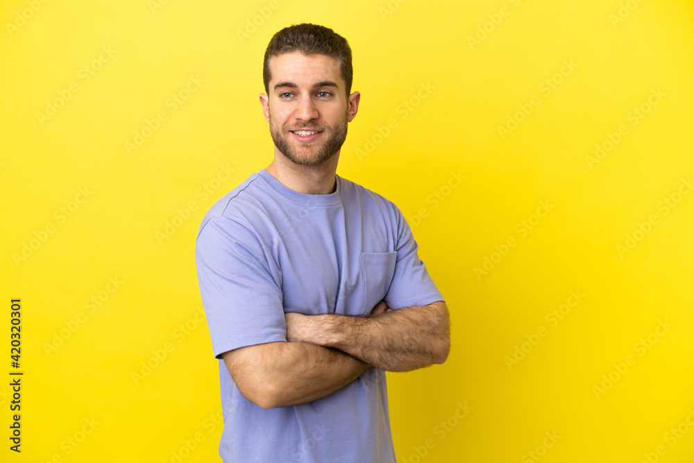 Handsome blonde man over isolated yellow background with arms crossed and happy