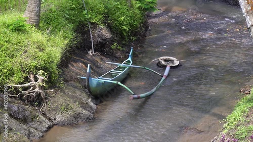 A blue wooden jukung (kano) is moored on the shore of a small river among a rice terrace in Bali.
