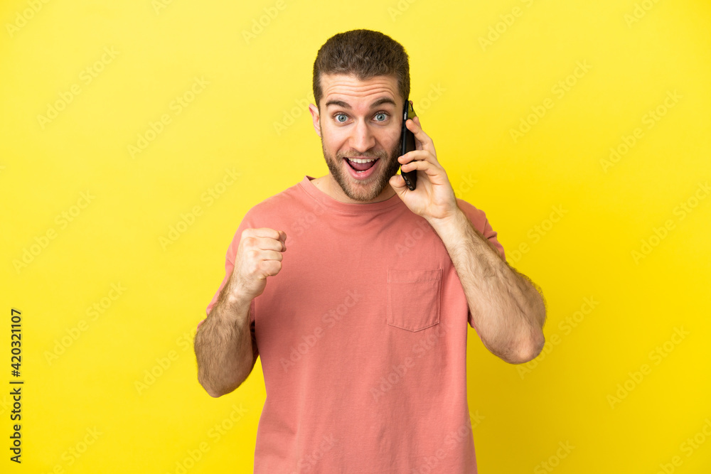 Handsome blonde man using mobile phone over isolated background celebrating a victory in winner position Handsome blonde man using mobile phone over isolated background celebrating a victory in winner position