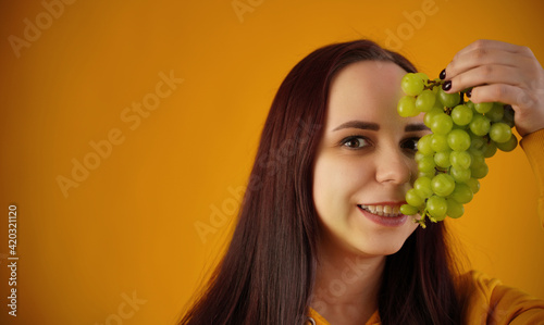 Portrait of young woman with green grapes on yellow background. Close up of pretty brunette closes eye of bunch of grapes.