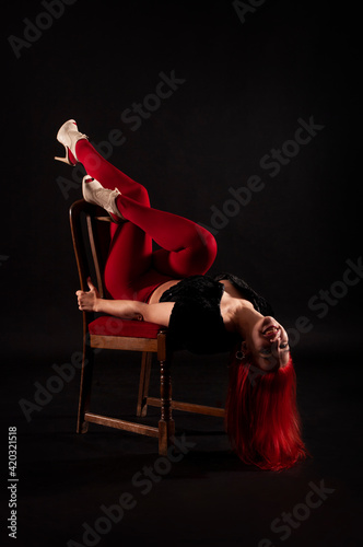 A girl with red hair, wearing red leggings poses on a chair on a dark background.