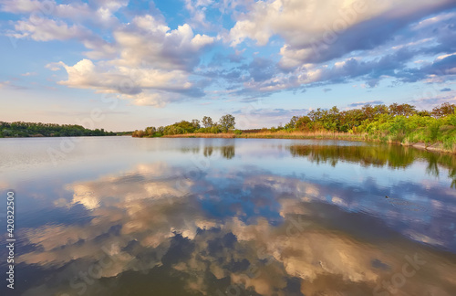 Beautiful river coast at sunset in summer. Colorful landscape with lake, green trees and grass, blue sky with multicolored clouds and orange sunlight reflected in water.