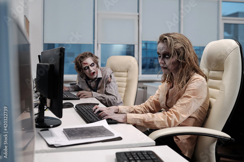 Young spooky zombie businesswoman sitting by desk in front of computer monitor and networking