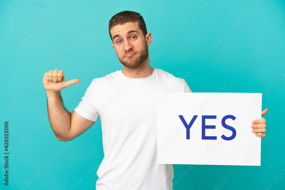 Handsome blonde man over isolated blue background holding a placard with text YES with proud gesture Handsome blonde man over isolated blue background holding a placard with text YES with proud gesture