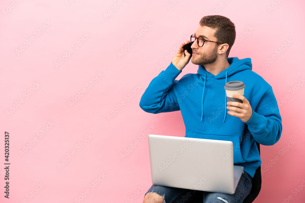 Young man sitting on a chair with laptop holding coffee to take away and a mobile Young man sitting on a chair with laptop holding coffee to take away and a mobile