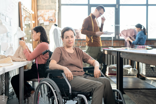 Young smiling seamstress in casualwear sitting in wheelchair against group of working colleagues