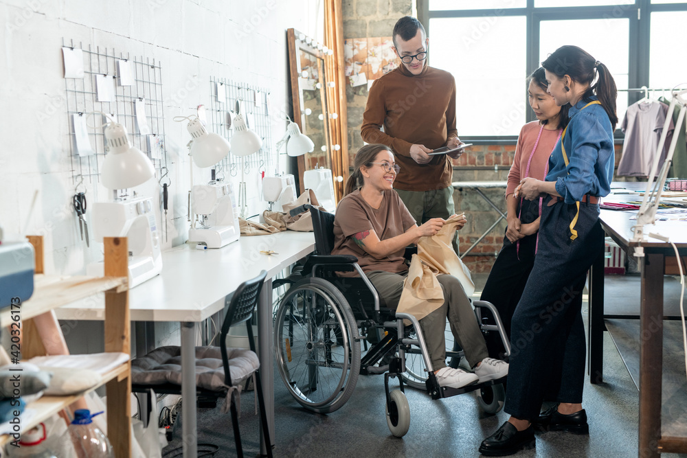 Group of young fashion designers surrounding disable seamtsress in wheelchair during discussion