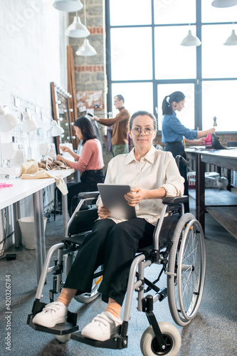 Young smiling seamstress with tablet sitting in wheelchair against group of colleagues in workshop